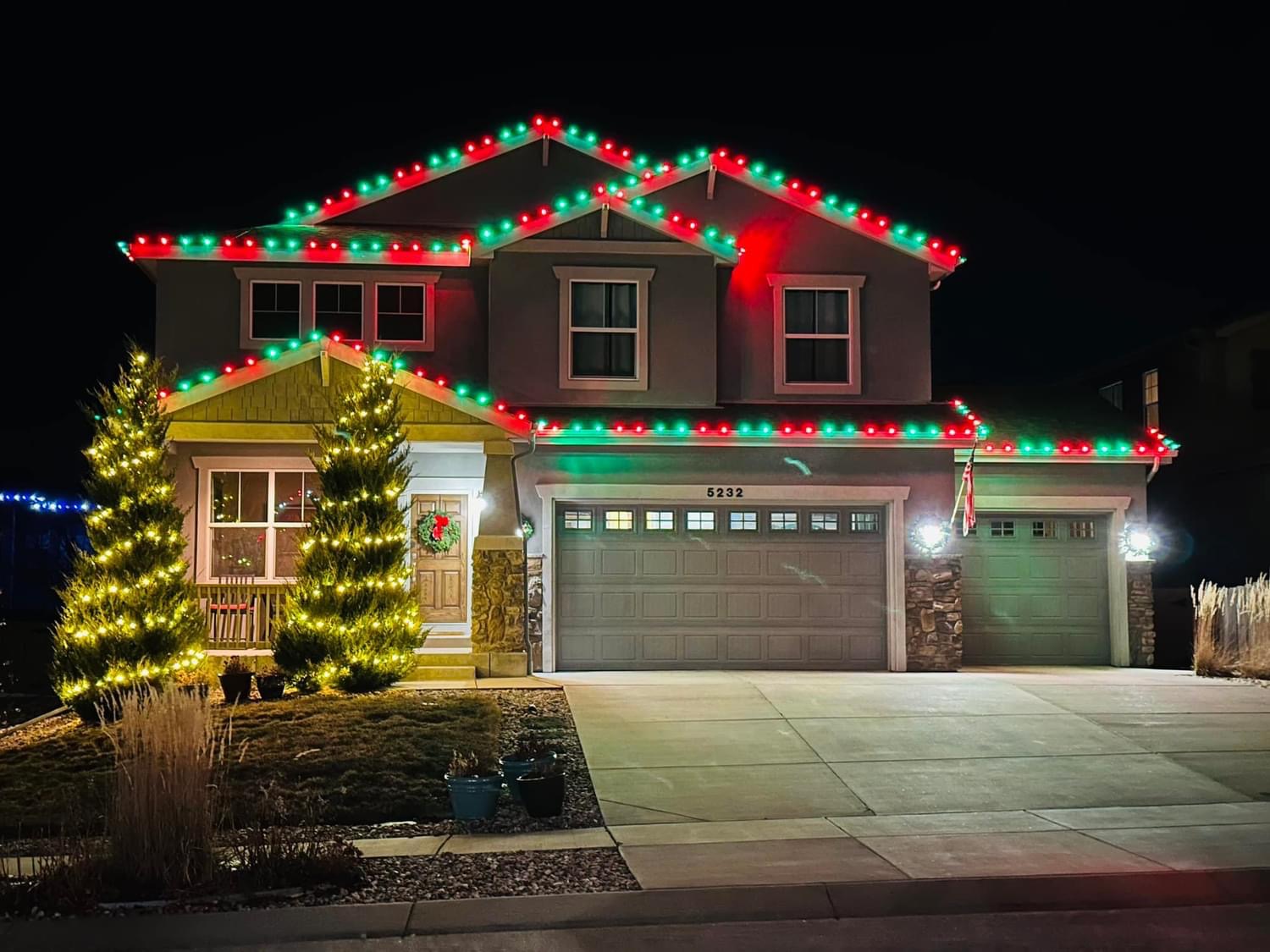 Red and green Christmas lighting on a two-story home with lit evergreens